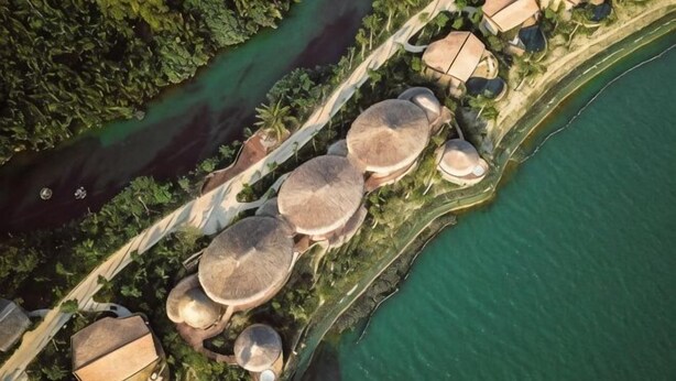 An aerial view of a coastal property with multiple round-roofed structures, a pathway, and a body of water. - Namia River Retreat - Wellness Inclusive Resort (Hoi An)