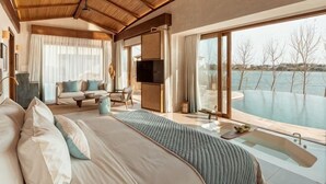 A bedroom with a large bed, a TV, a sofa, a dining table, and a view of a pool. - Namia River Retreat - Wellness Inclusive Resort (Hoi An)