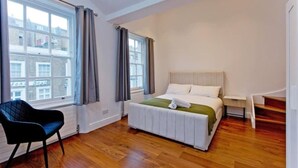 Ein Schlafzimmer mit einem Bett, einem Sessel, einem Nachttisch und einem Fenster mit Vorhängen.