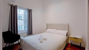 Ein Schlafzimmer mit Bett, Sessel, Nachttisch und einem Fenster mit Vorhängen.