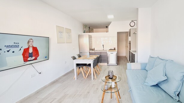A modern living room with a TV displaying a news broadcast, a dining area with a table and chairs, a light blue sofa, and a kitchen in the background. - Playitas Salema Apartment (Las Playitas)