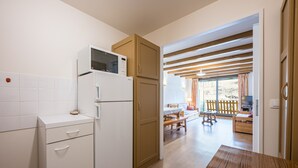Une cuisine compacte équipée d’un four à micro-ondes, d’un réfrigérateur et de meubles en bois. Une table à manger se trouve au premier plan, et un balcon avec une balustrade en bois est visible à l’arrière-plan.
