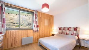 Une chambre avec un mur en bois, un lit, une fenêtre avec des rideaux, une table de chevet avec une lampe et une commode.