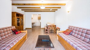 Un salon avec deux canapés, une table basse, des poutres en bois, un meuble en bois et, au fond, une cuisine.