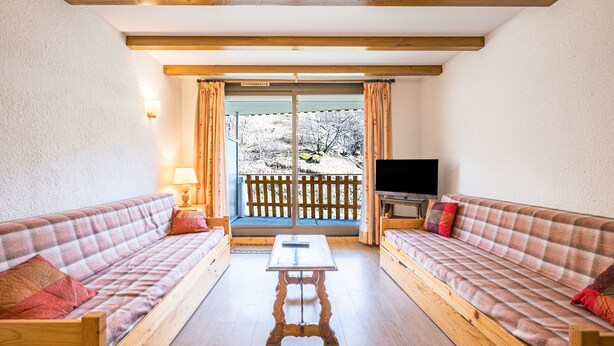 Une pièce comprenant deux canapés en bois, une table en bois, un téléviseur et un balcon avec vue sur les arbres.