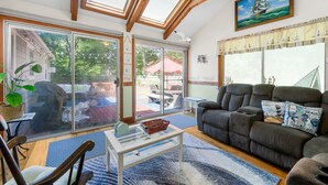 A cozy living room with a large gray sofa, a glass coffee table, and a view of the outdoors through a sliding glass door. - Life's Better at Marshside Madness! (West Yarmouth)