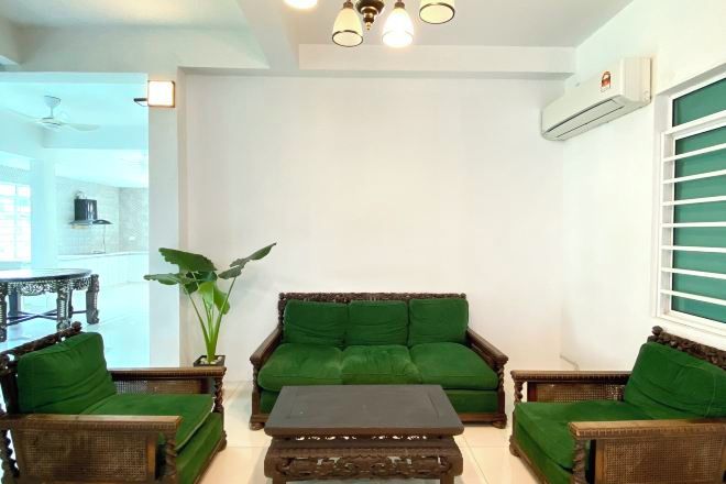 A living room with a green sofa, two wooden armchairs, and a coffee table. There is a ceiling fan, an air conditioning unit, and a window with blinds.