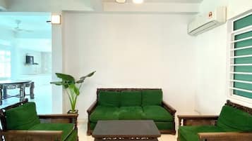 A living room with a green sofa, two wooden armchairs, and a coffee table. There is a ceiling fan, an air conditioning unit, and a window with blinds.