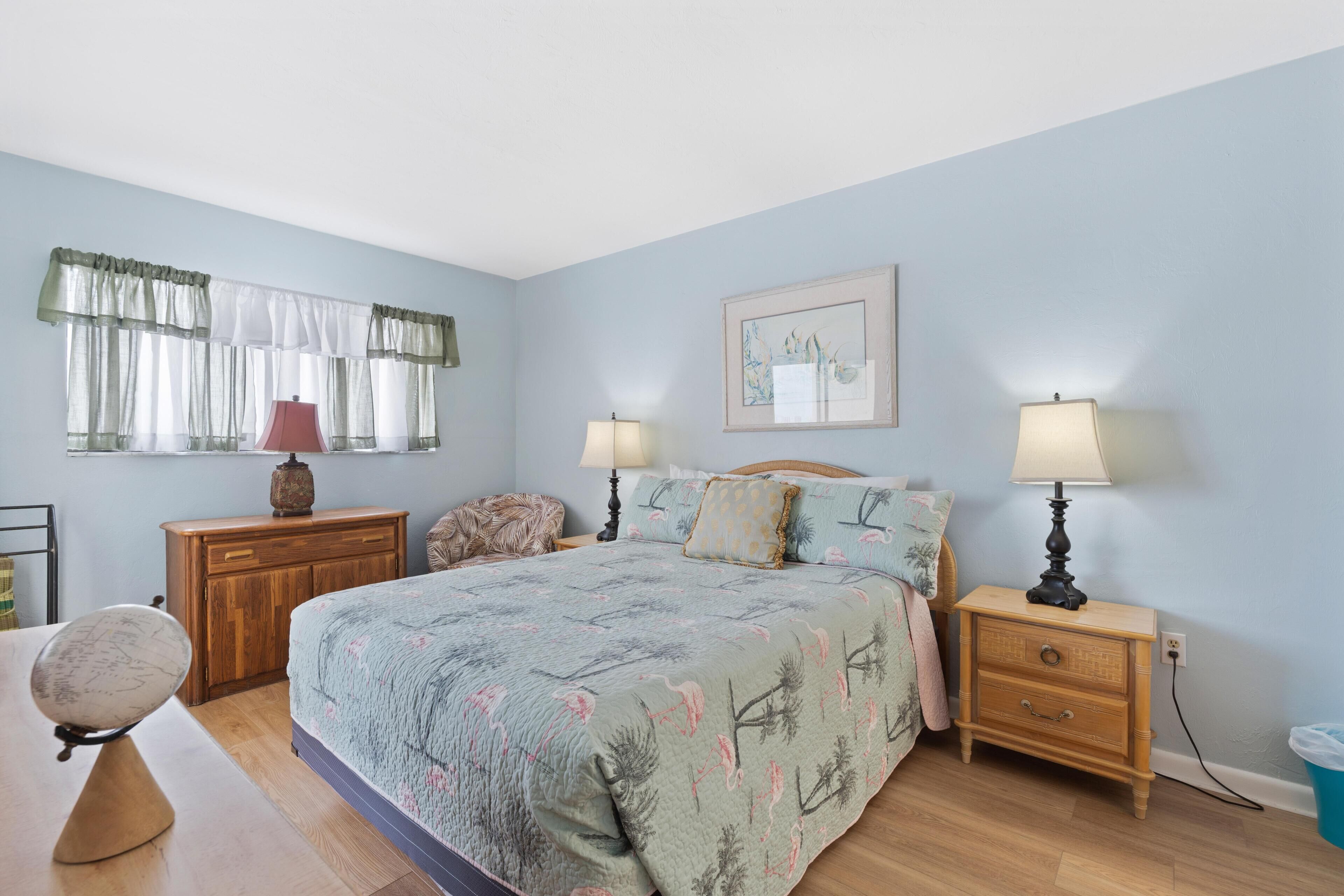 A bedroom with a bed, two bedside tables, a nightstand, a wooden dresser, a lamp, a framed picture, and a window with curtains.