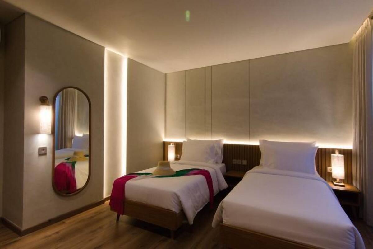 A hotel room with two beds, a round mirror, bedside lamps, and a wooden floor.