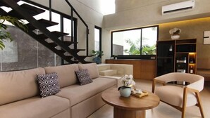 A modern living room with a sofa, coffee table, and chair, a staircase, and a kitchen area with a large window. - Umahan Sanur (Bali)