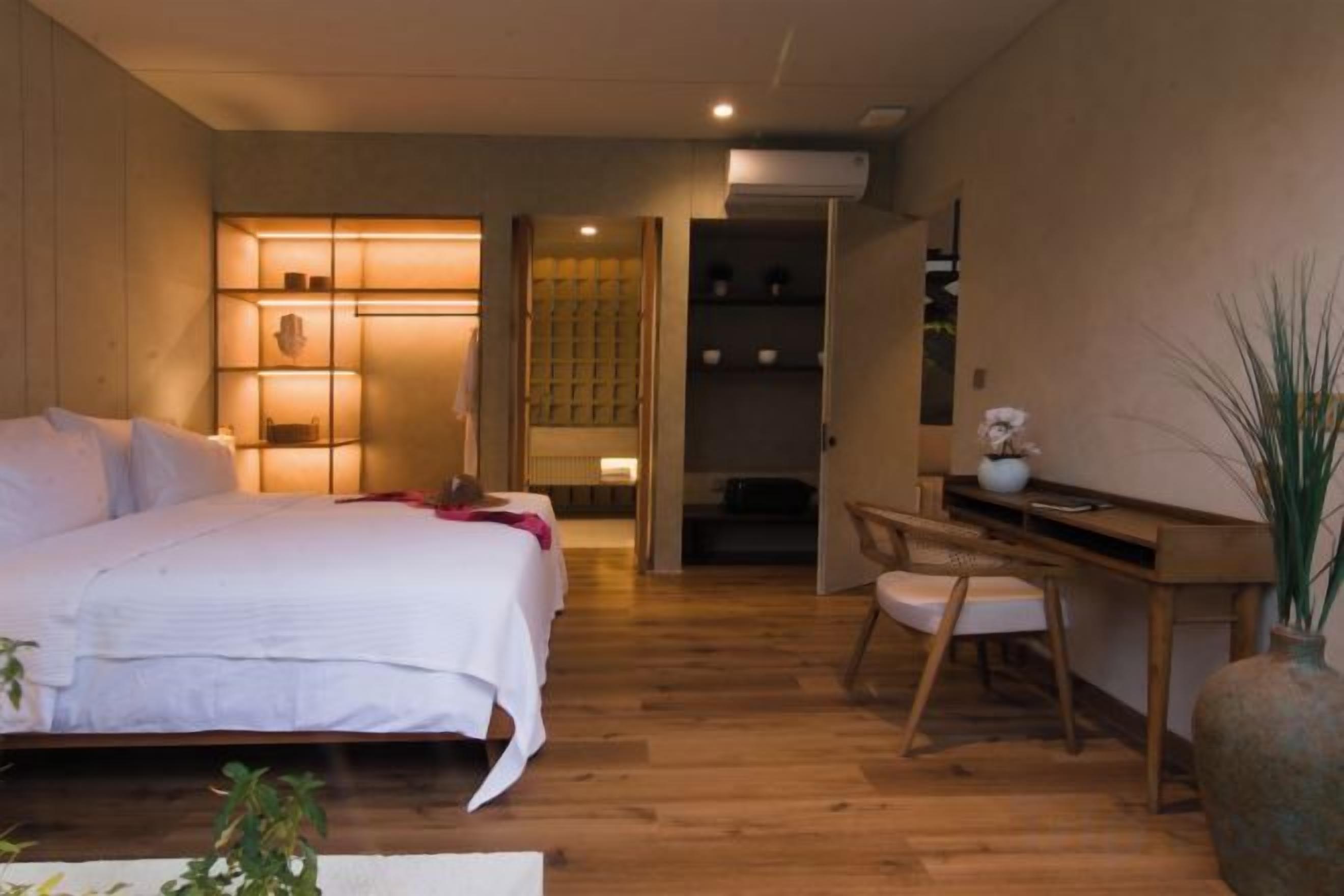 A bedroom with a bed, bedside table, chair, and a desk with a chair. There is a plant and a vase on the desk.