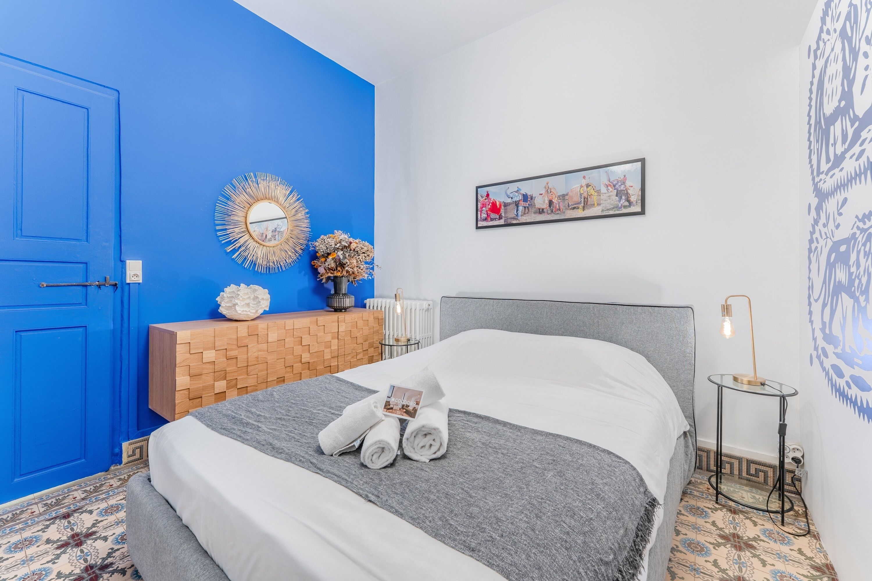 Una camera d'albergo con una porta blu, un letto con lenzuola bianche, un tavolino con una lampada e uno specchio fissato alla parete.