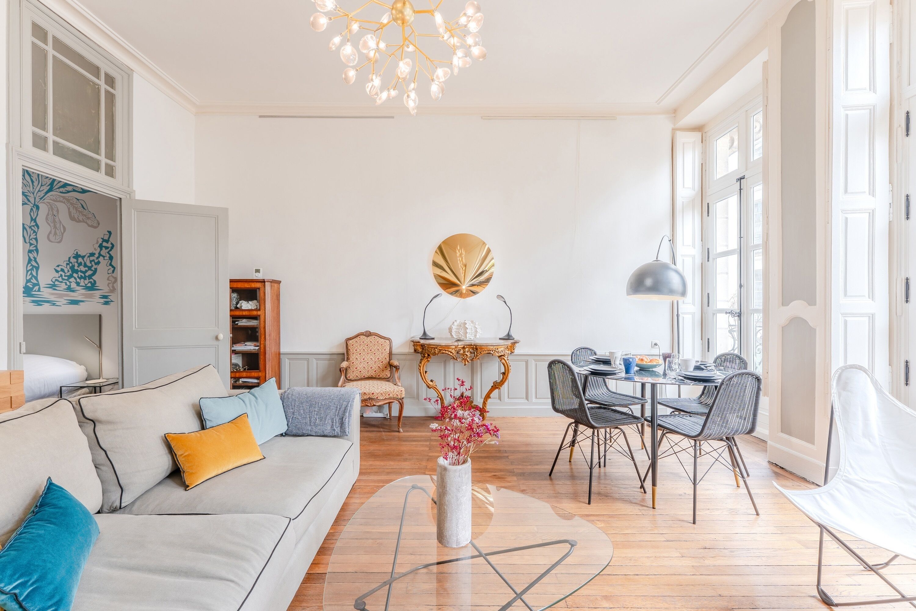 Uma sala de estar espaçosa com sofá, mesa de jantar e cadeiras. Há um candelabro, um vaso com flores e uma grande janela que ilumina naturalmente o ambiente.