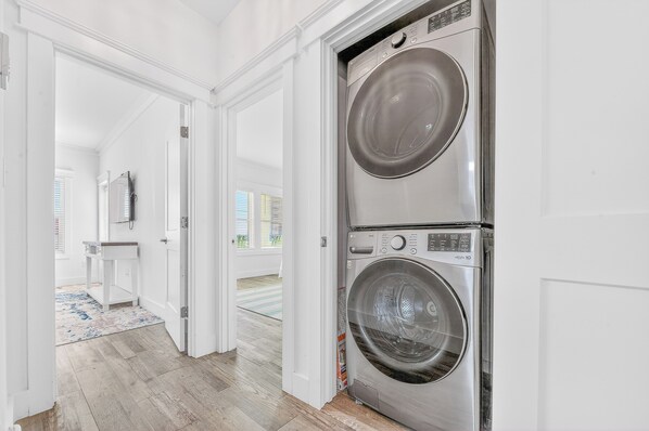 A laundry room with two stacked front-loading washing machines, a white door, and a view into a bathroom with a mirror and a sink. - Lux 6BR - Heated Pool | Sleeps 22 | 1 min to beach (Gulf Shores)