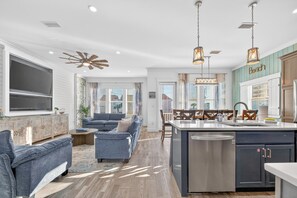 A spacious living room with a large TV, a sofa, and a dining area with a kitchen island. - Lux 6BR - Heated Pool | Sleeps 22 | 1 min to beach (Gulf Shores)