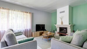 Ein Wohnzimmer mit einer Sofagarnitur, einem Fernseher, einem Kamin und einem Fenster mit Vorhängen.