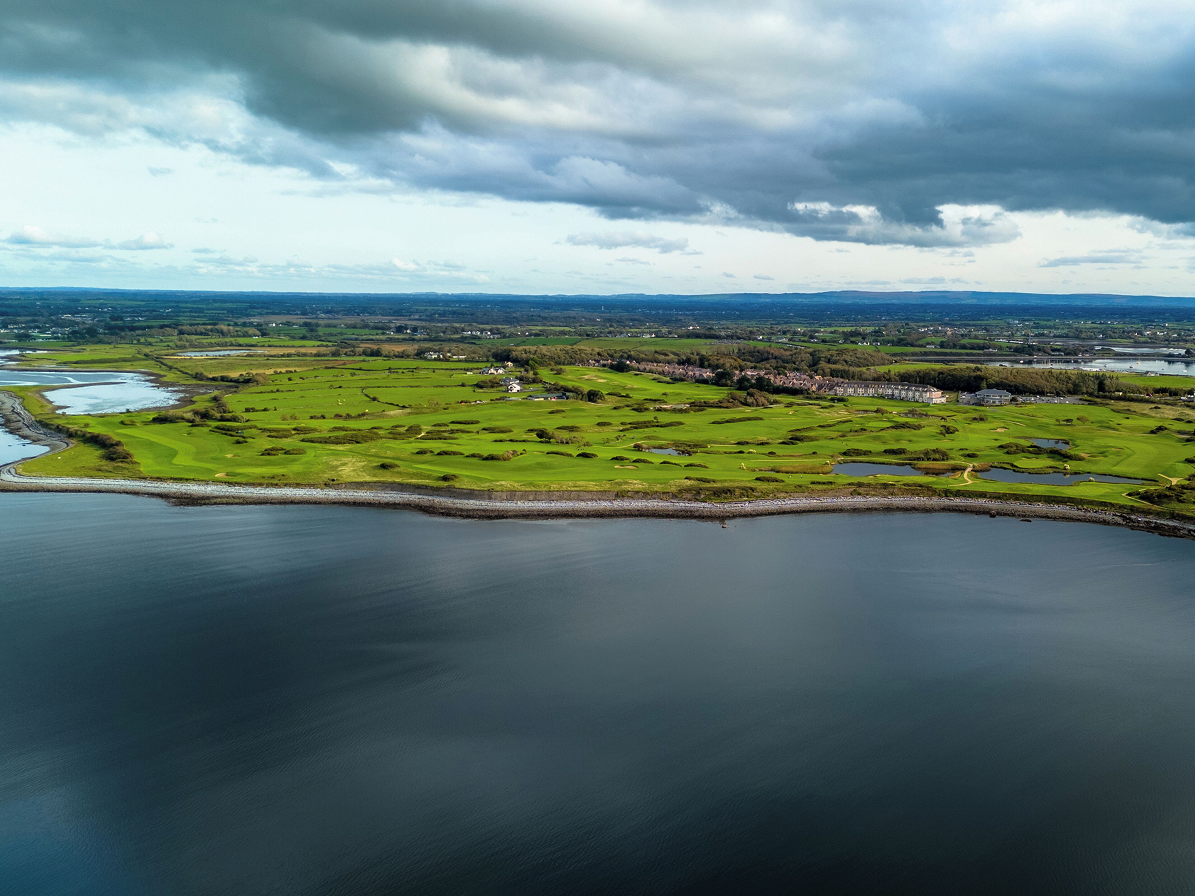An aerial view of a golf course with a body of water in the foreground, surrounded by green fields and a few buildings.