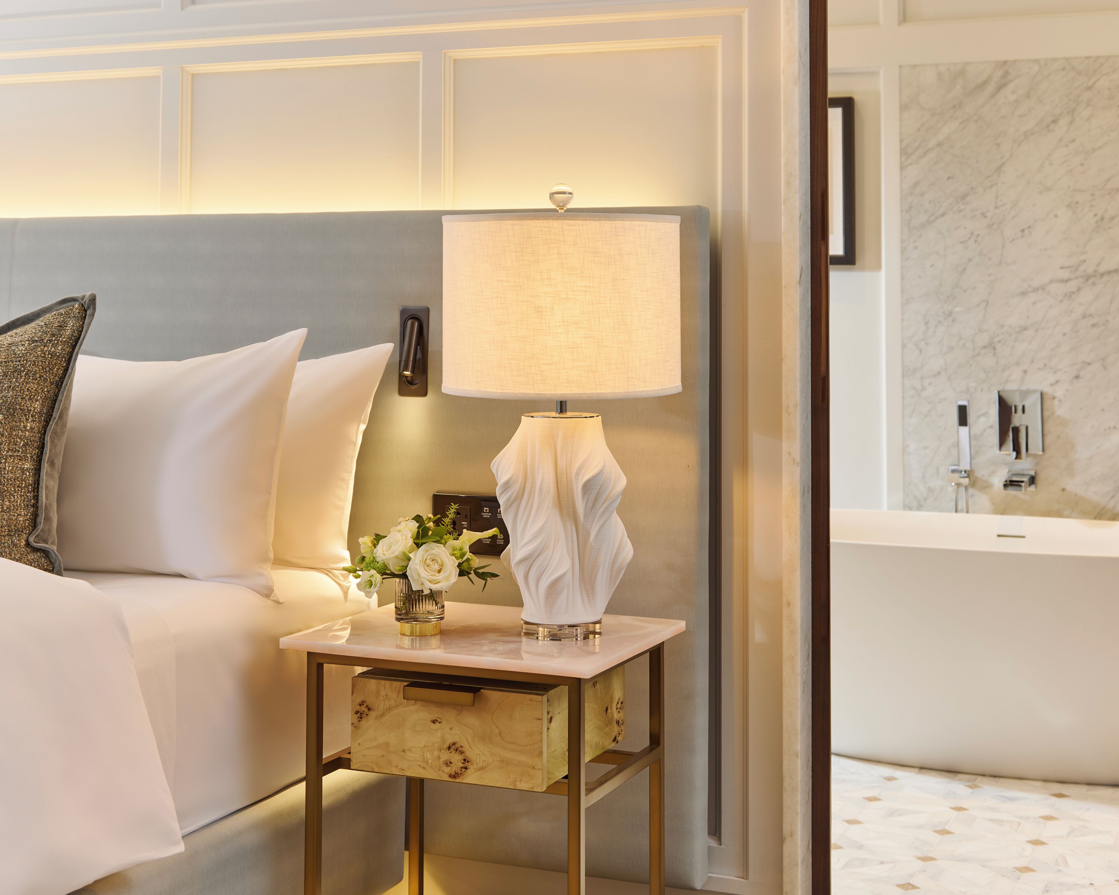 Habitación de hotel con cama, mesita de noche con lámpara, jarra con flores y vista al baño con bañera y pared de mármol.