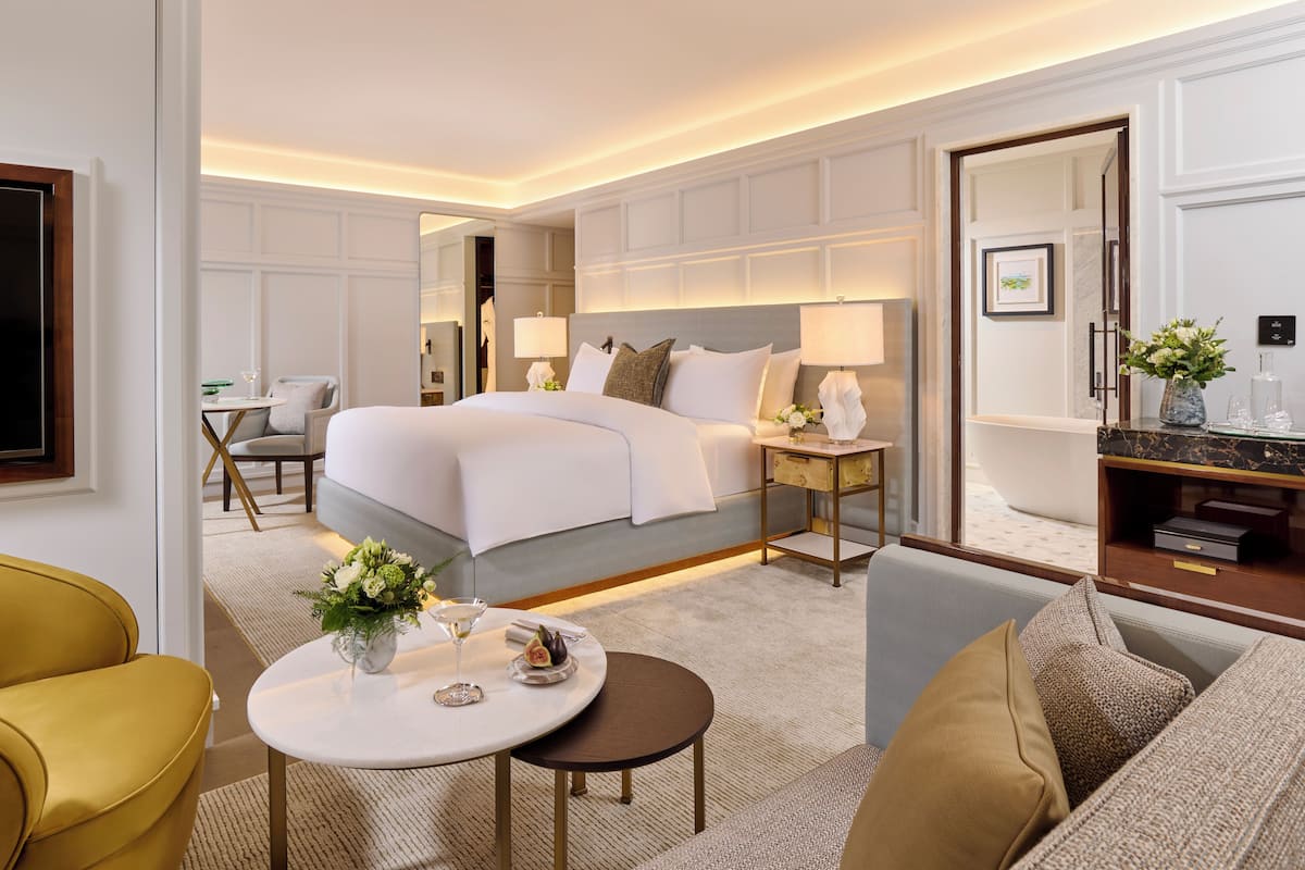 Habitación de hotel con una cama grande, un área de descanso con un sofá y sillones, una mesita redonda con un florero y una copa, y un tocador con espejo y artículos de aseo.