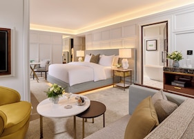 Habitación de hotel con una cama grande, un área de descanso con un sofá y sillones, una mesita redonda con un florero y una copa, y un tocador con espejo y artículos de aseo.
