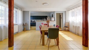 Un comedor con una mesa y sillas de madera, una cocina al fondo y amplios ventanales con cortinas.