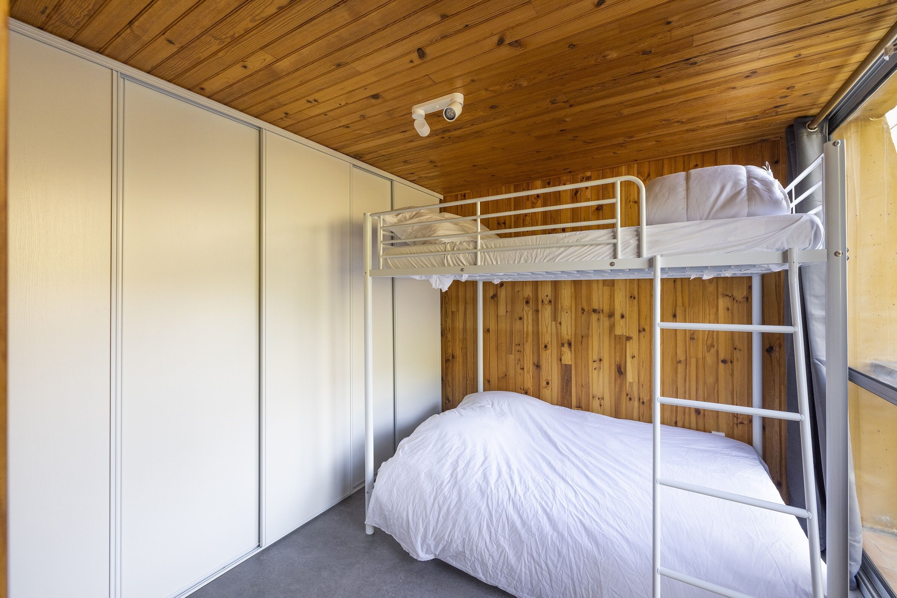 A bunk bed room with a wooden ceiling, a white wardrobe, and a security camera.