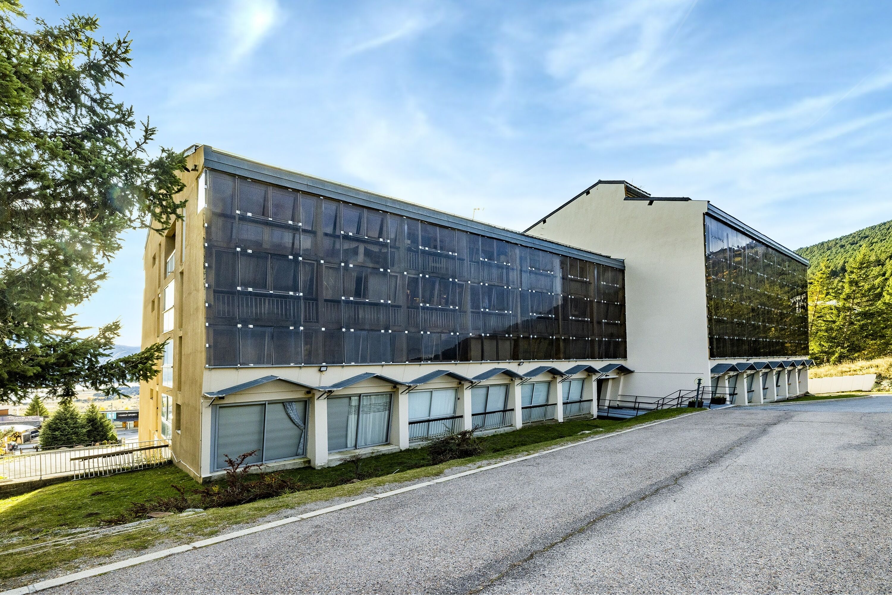 Een modern gebouw met grote ramen en een plat dak, omringd door groen en een heldere lucht.