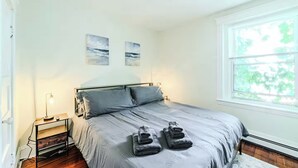 Une chambre à coucher comprenant un lit, deux œuvres d’art murales, une table de chevet avec une lampe et une fenêtre donnant sur un paysage verdoyant.