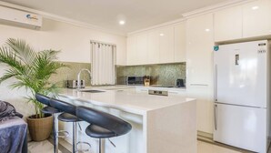 A modern kitchen with white cabinets, a large island with bar stools, a refrigerator, and a potted plant. - Boardwalk Bliss Explore the Lively Mindarie Marina (Mindarie)