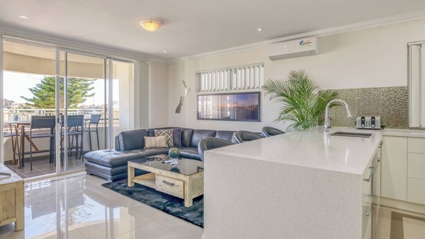 A modern living room with a sofa, coffee table, and a dining area with chairs. There is a kitchen area with a sink and a large window. - Boardwalk Bliss Explore the Lively Mindarie Marina (Mindarie)