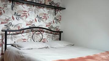 A bedroom with a bed, a wooden floor, a floral wallpaper, and a shelf with a vase.