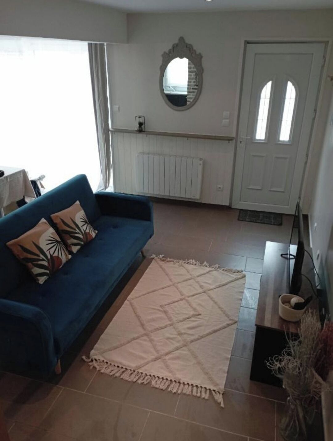 Una sala de estar con sofá azul, cojín decorativo estampado, alfombra color beige, espejo en la pared y puerta blanca.