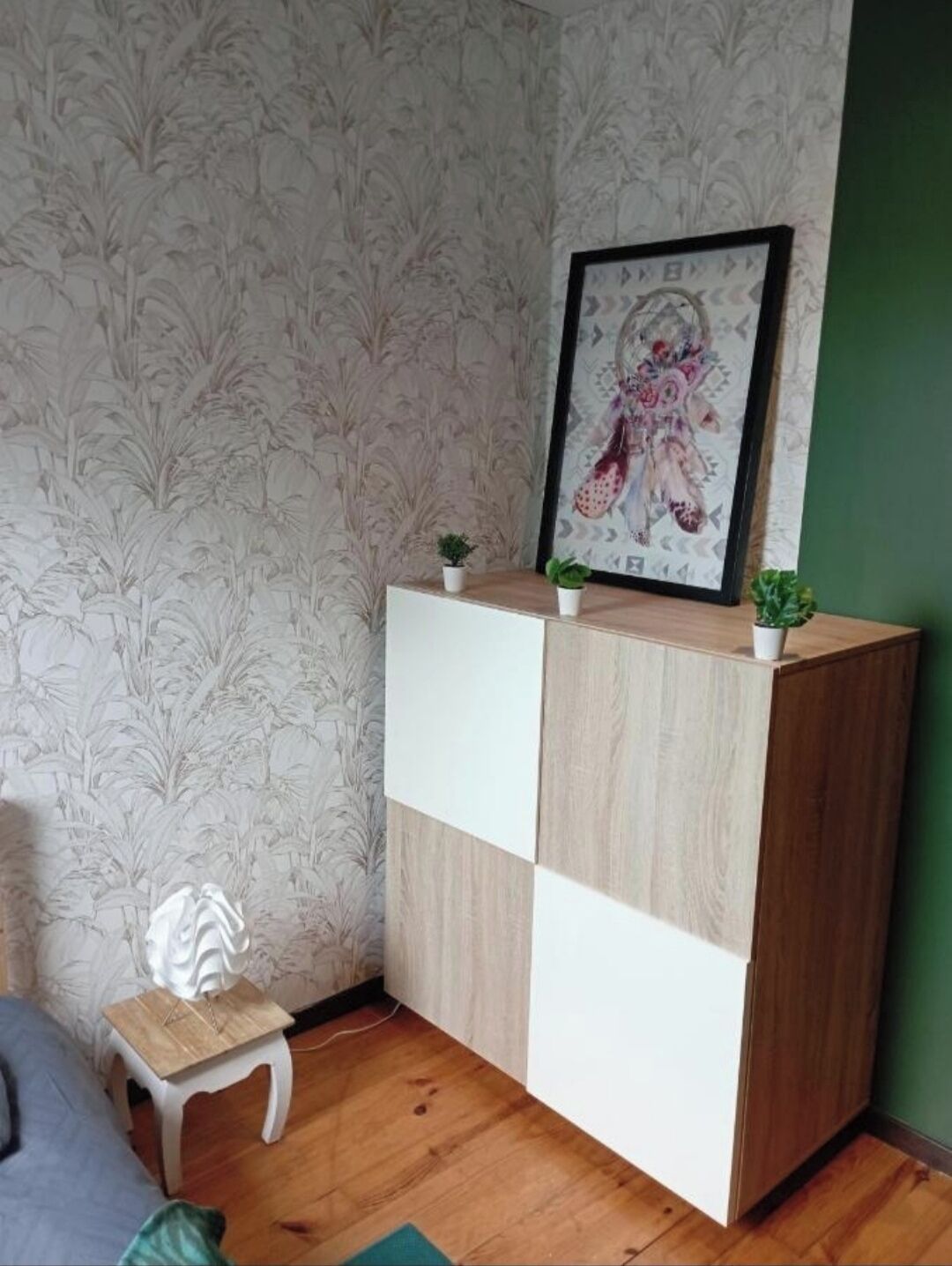 A corner room with patterned wallpaper, a wooden cabinet with a picture frame, a small white stool, and a green wall.