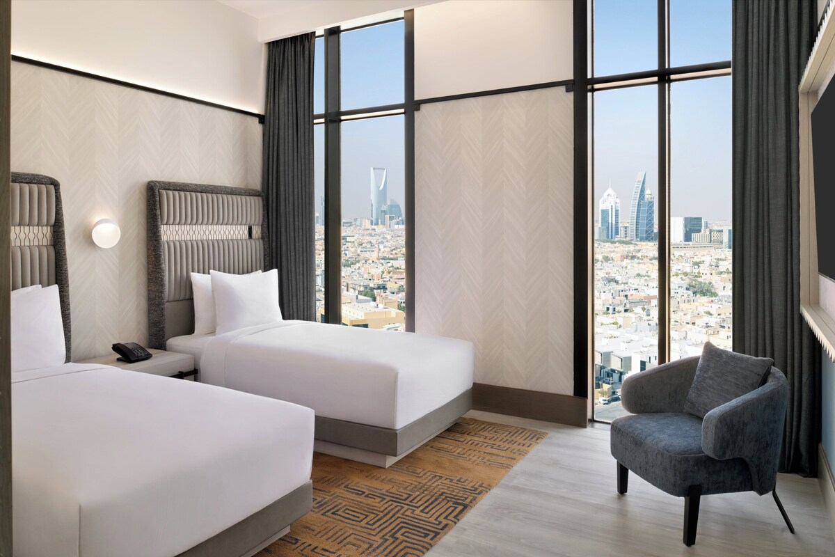A hotel room with two beds, a chair, and a large window offering a city view.