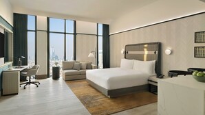 Quarto de hotel moderno com uma cama grande, escrivaninha, sofá e vista para a cidade através de amplas janelas.