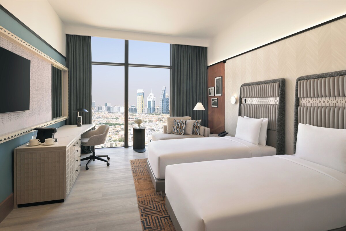 A hotel room with two beds, a desk, a TV, and a large window offering a city view.