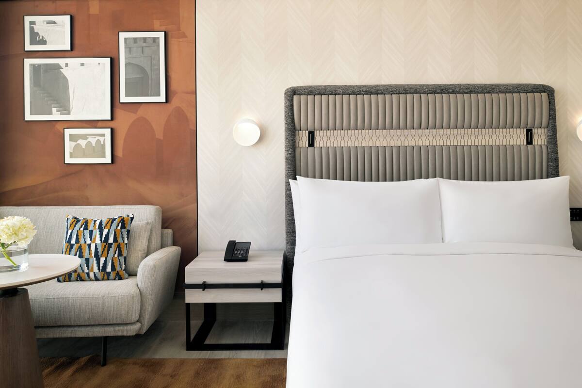 Habitación de hotel con una cama grande, una mesita de noche con teléfono, una silla con cojín estampado y una pared con cuadros.