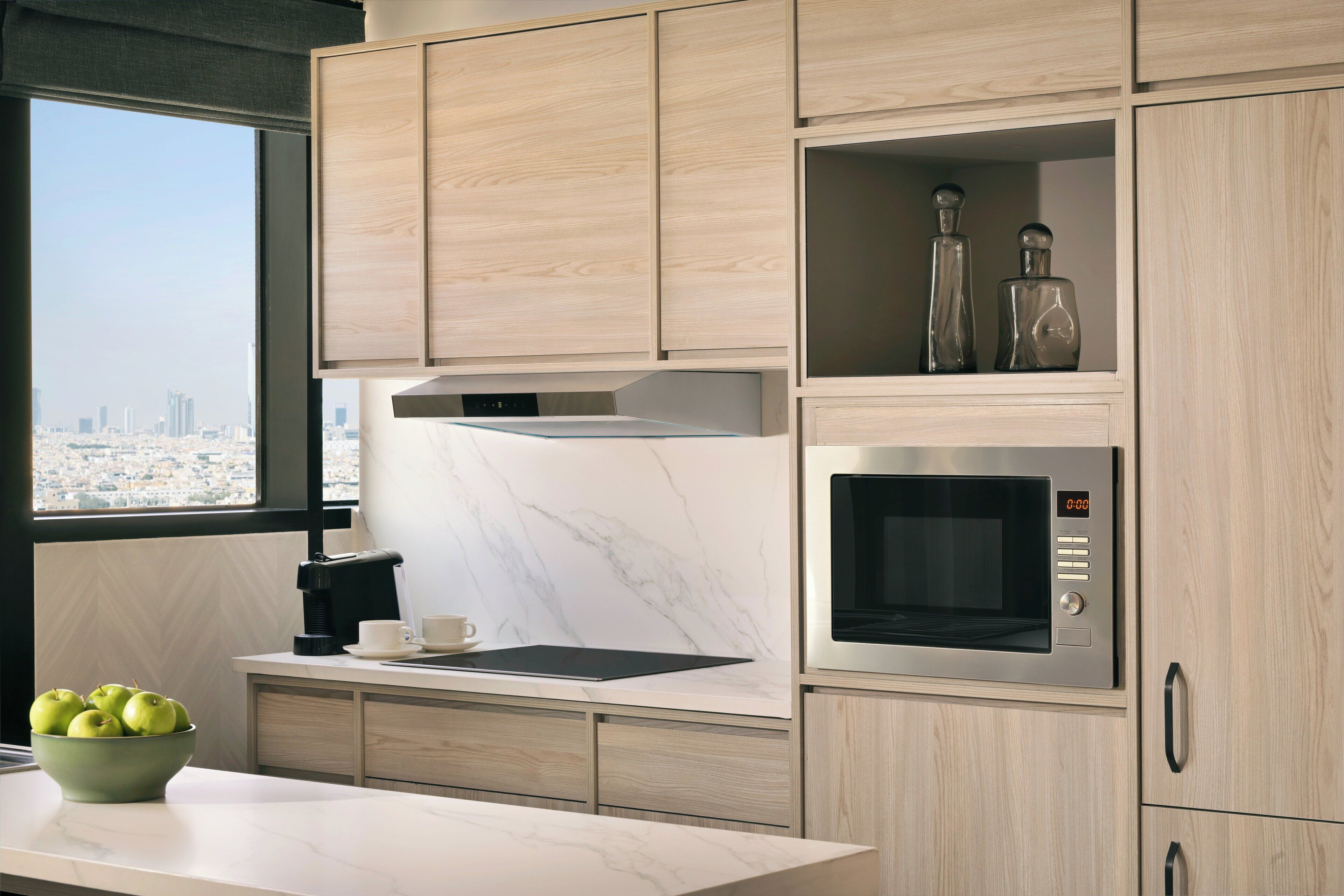 A modern kitchen with wooden cabinets, a built-in microwave, and a view of the city through a large window.