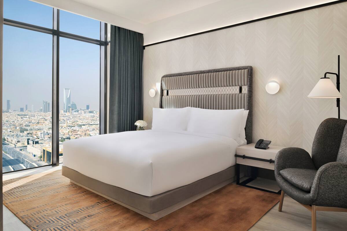 Habitación de hotel moderna con una cama grande, una mesita de noche, una silla y vistas a la ciudad a través de amplios ventanales.