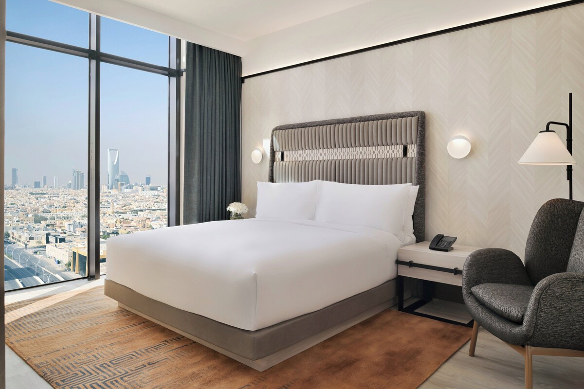 A modern hotel room with a large bed, a nightstand, a chair, and a city view through large windows.