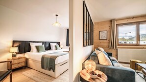 A modern hotel room with a large bed, a sitting area with a sofa and a table, and a view of the outside through a large window. - Mimosa–Apartment near piste, mountain-view balcony (Les Gets)