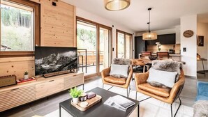 A modern living room with a large TV, wooden furniture, and a dining area in the background. - Mimosa–Apartment near piste, mountain-view balcony (Les Gets)