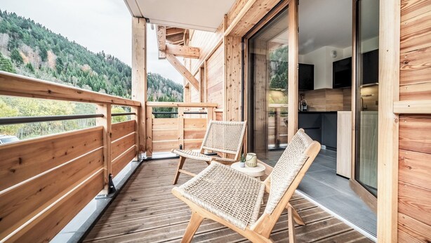 A wooden balcony with two wicker chairs and a small table, overlooking a forested hillside. - Mimosa–Apartment near piste, mountain-view balcony (Les Gets)