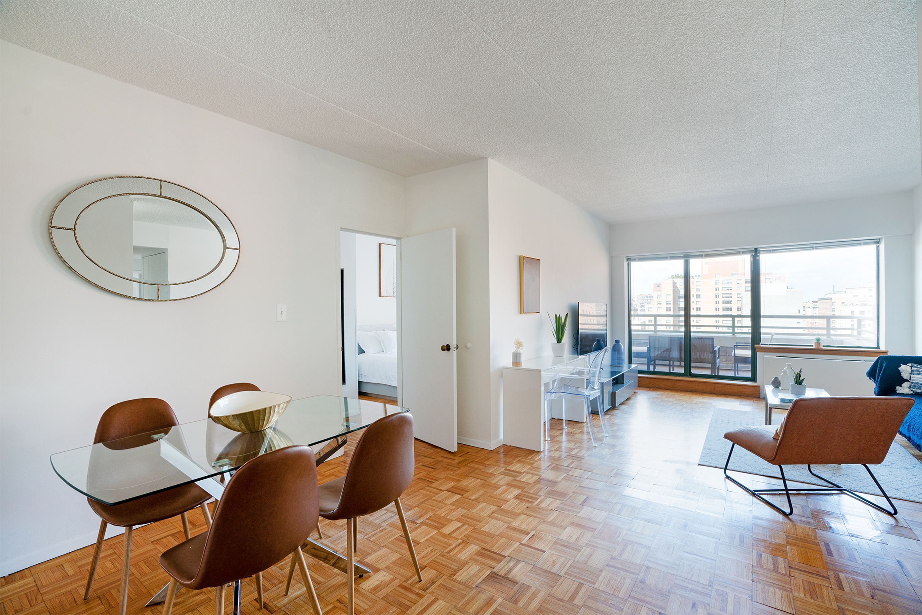 A modern living room with a glass dining table, brown chairs, a round mirror, and a large window offering a city view.