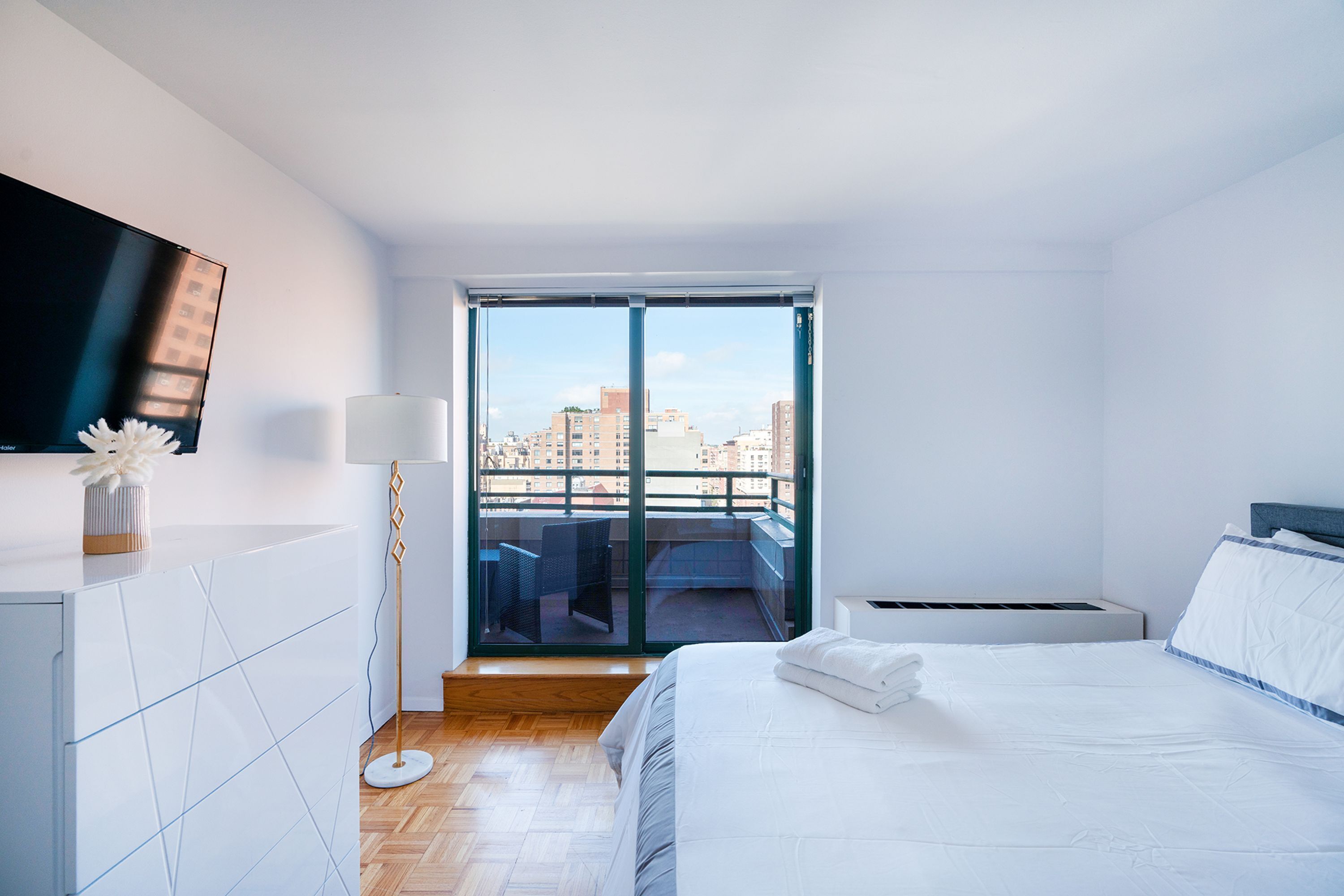 A modern hotel room with a large bed, a flat-screen TV, a balcony with a city view, and a floor lamp.