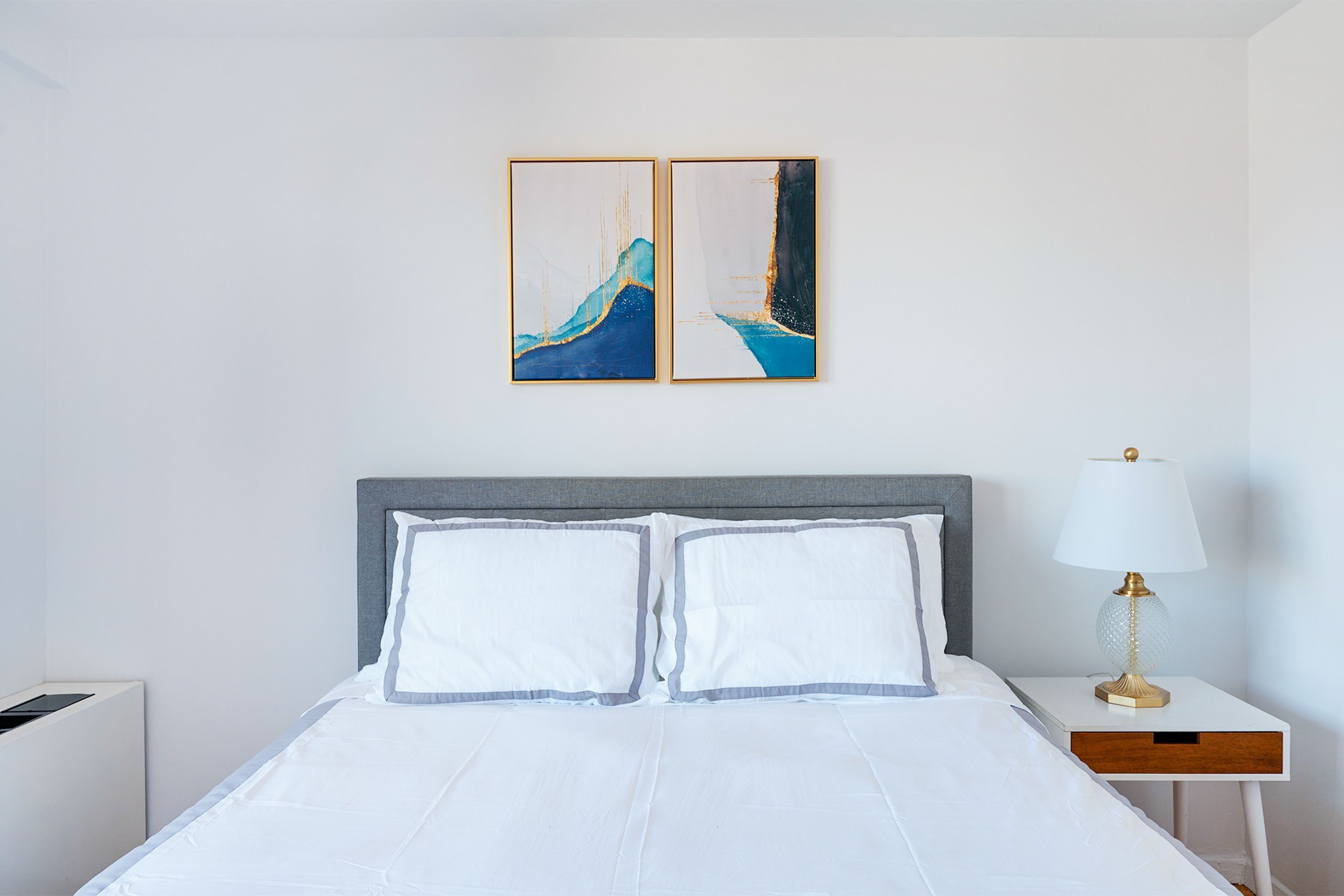 A hotel room with a bed, a nightstand with a lamp, and two wall art pieces.
