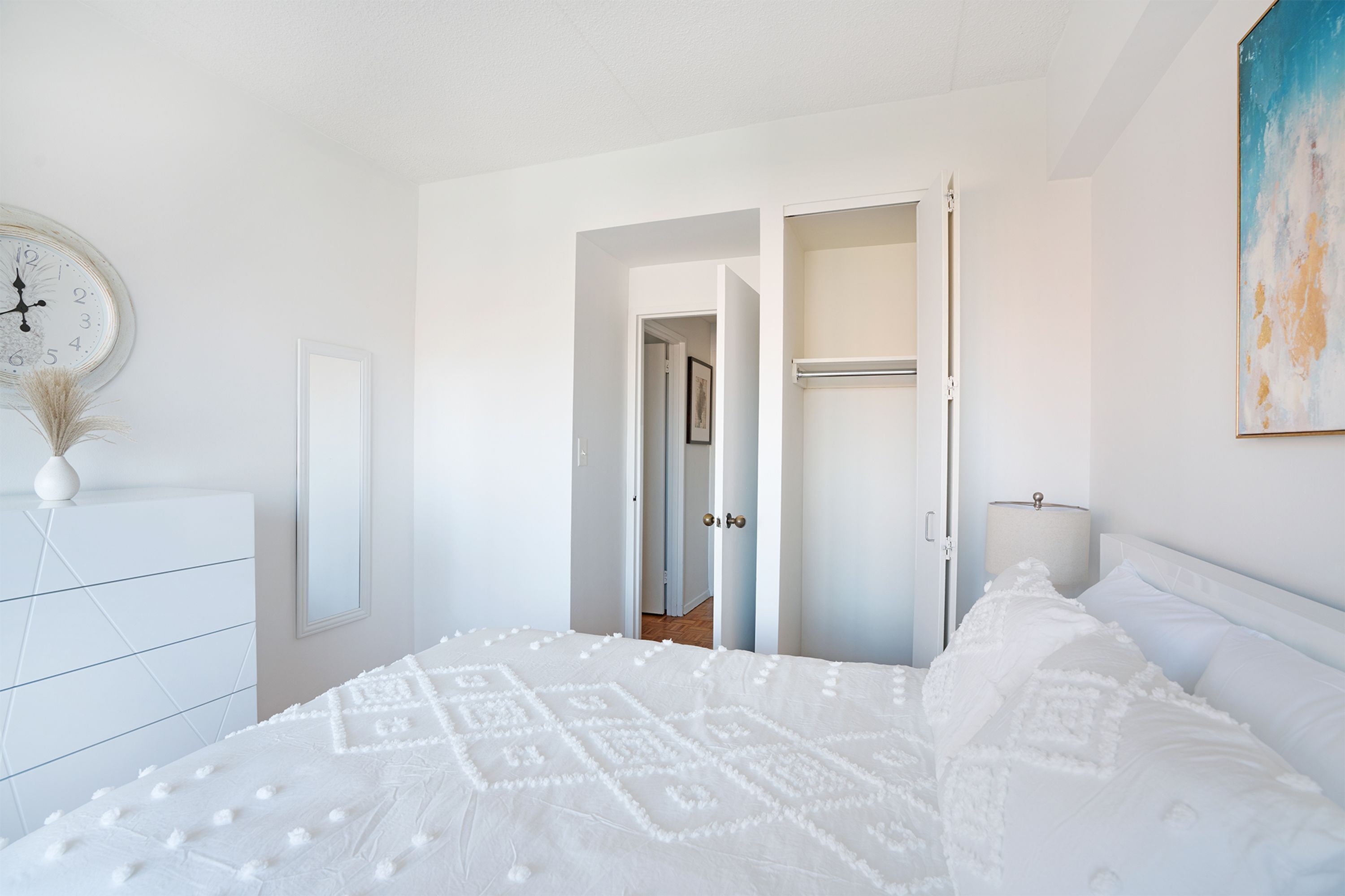 A bedroom with a large bed, a dresser, a clock, and a door leading to another room.