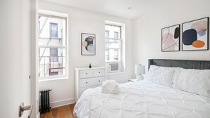A bedroom with a large bed, two windows, a radiator, a white dresser, and abstract wall art. - 1 BR Gem in Prime Location of NYC (New York)