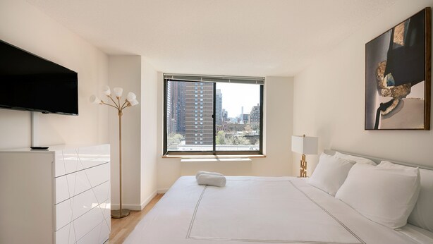 A modern hotel room with a large bed, a flat-screen TV, a floor lamp, and a city view through the window. - Stunning Apartment for 4 2 Bedrooms 2 Bath (New York)